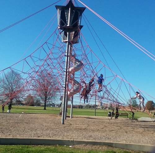 Children play on a large red climbing structure in a park under a clear blue sky.