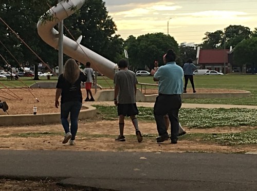 Three people walking in a park near a playground, with a slide and trees in the background.
