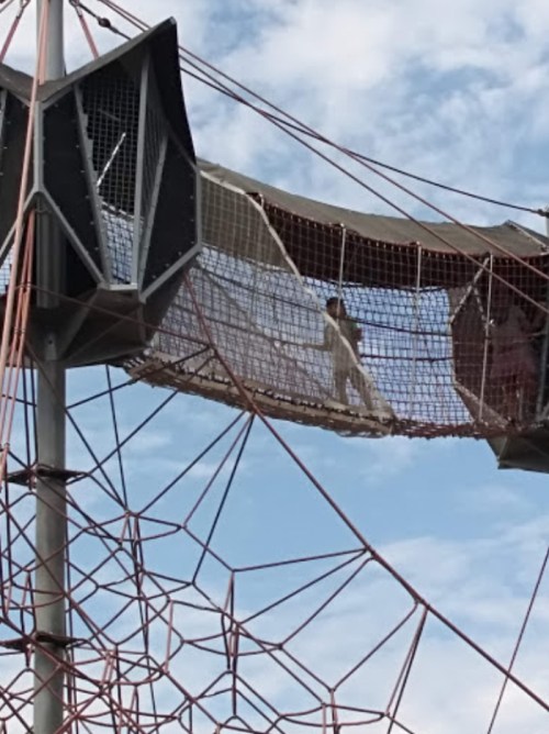 A person walks on a suspended net structure against a blue sky, surrounded by ropes and climbing elements.