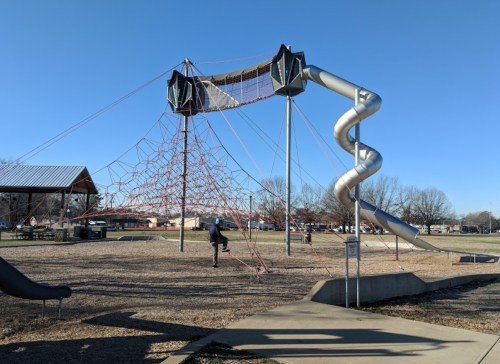 A playground featuring a climbing structure, a spiral slide, and a person climbing the net under a clear blue sky.