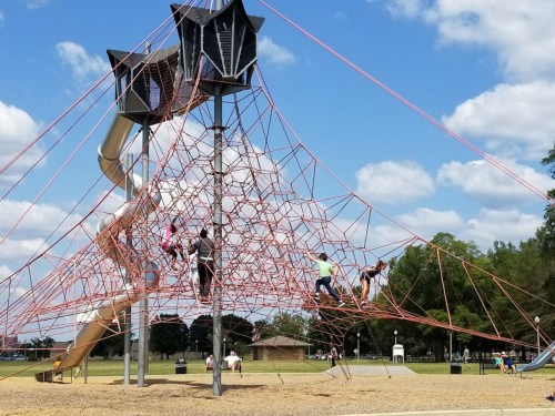 Children climbing on a large rope structure at a park, with a slide and blue sky in the background.