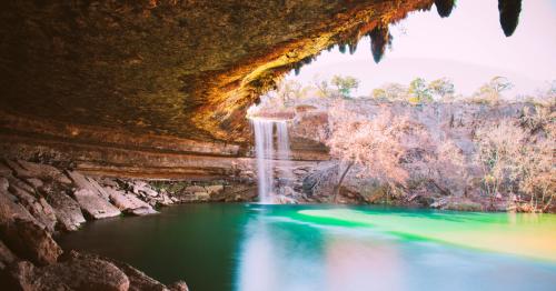 A serene waterfall cascades into a turquoise pool, surrounded by rocky cliffs and lush trees under a bright sky.