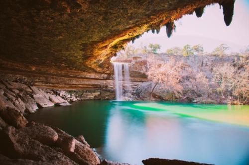 A serene waterfall cascades into a turquoise pool, framed by rocky cliffs and lush trees in a tranquil setting.