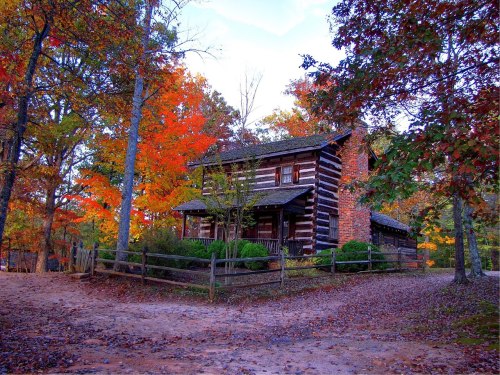 A rustic log cabin surrounded by vibrant autumn foliage and a dirt path, set in a serene forested area.