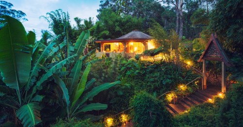 A warm, lighted tropical villa at night surrounded by dense jungle plants and large banana leaves in Hawaii.