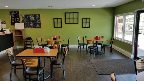 A cozy café interior with green walls, empty tables, and a chalkboard menu. Large windows let in natural light.