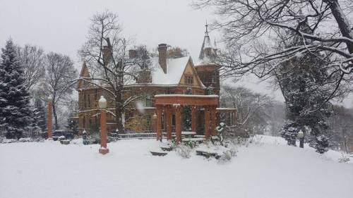 A snow-covered mansion surrounded by trees and a brick archway, creating a serene winter scene.