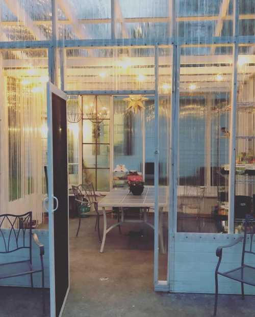 A cozy greenhouse with transparent walls, featuring a table and chairs, illuminated by warm lights.