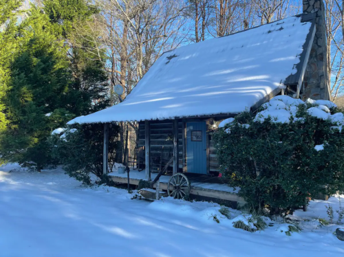 A cozy log cabin covered in snow, surrounded by trees, with a blue door and a rustic wheel in the front.