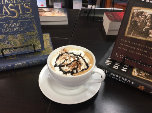 A cup of coffee topped with whipped cream and chocolate drizzle, surrounded by books on a table.