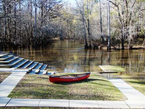 A red canoe rests on the grass by a calm, reflective river surrounded by trees and a set of stone steps.