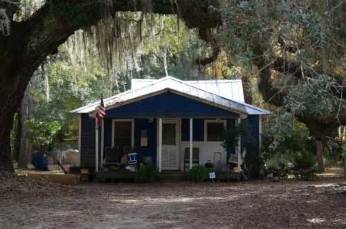 A blue house with a white roof, surrounded by trees and Spanish moss, featuring an American flag on the porch.