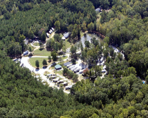 Aerial view of a wooded campground with RVs and cabins surrounded by trees and a winding road.