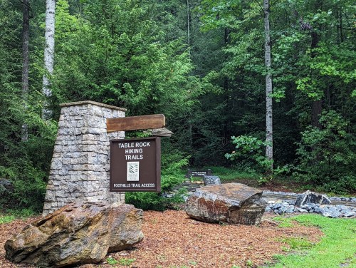 Sign for Table Rock Hiking Trails surrounded by trees and rocks, indicating trail access in a lush forest setting.