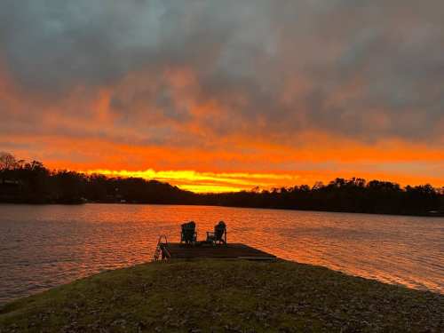 A serene lake at sunset, with vibrant orange and yellow hues in the sky and two chairs on a dock.