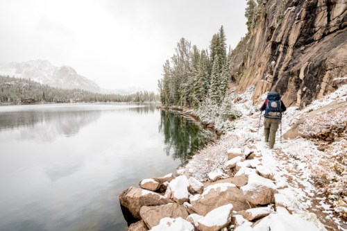 A hiker walks along a snowy lakeside trail, surrounded by trees and mountains under a cloudy sky.