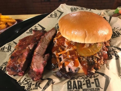 A plate featuring a BBQ sandwich with pulled meat, pickles, and sauce, alongside two slices of smoked brisket.