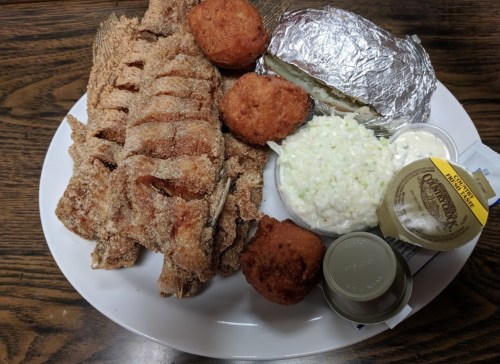 A plate of fried fish, hushpuppies, coleslaw, and a baked potato wrapped in foil on a wooden table.