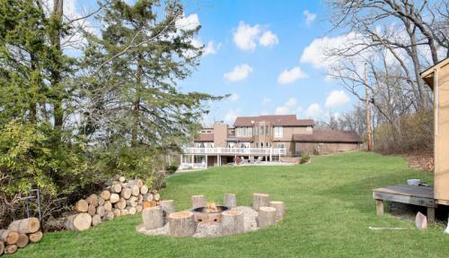 A spacious backyard with a fire pit, logs stacked nearby, and a modern house in the background under a blue sky.