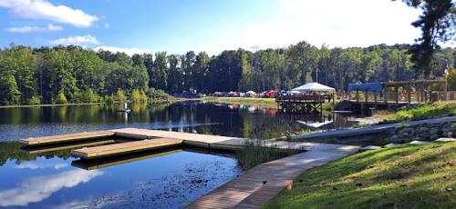 A serene lake scene with wooden docks, lush greenery, and tents set up along the shore under a clear blue sky.
