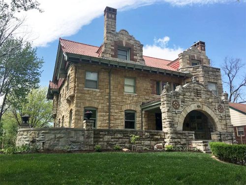 Historic stone house with a red tile roof, decorative archway, and lush green lawn under a blue sky.