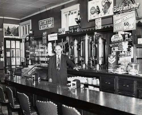 A man stands behind a bar with vintage decor, featuring signs for drinks and food, in a classic diner setting.