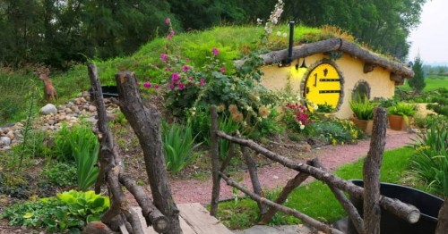 A whimsical earth-sheltered home in Nebraska with a round yellow door, grass roof, and a rustic branch fence.
