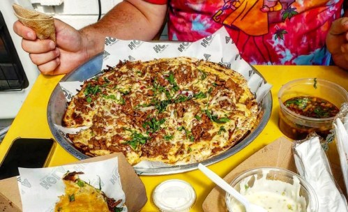 A person holds a cone while sitting at a table with a large pizza topped with meat and herbs, alongside dipping sauces.