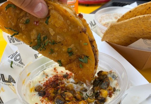 A hand holds a crispy taco above a bowl of creamy corn dip, garnished with herbs and spices.