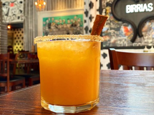 A vibrant orange cocktail with a sugared rim and a cinnamon stick, set on a wooden table in a stylish restaurant.