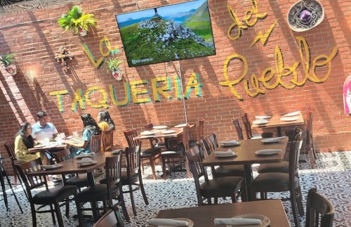 Interior of a taqueria with a TV, brick walls, and tables. People are dining and colorful decor is visible.
