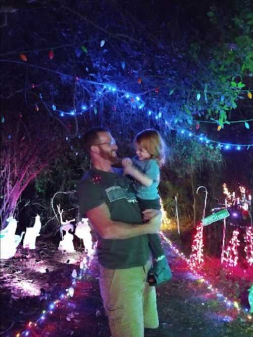 A man holds a child in a festive, brightly lit outdoor setting with colorful lights and decorations.