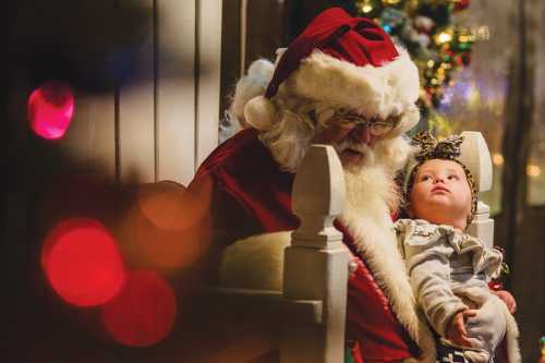 Santa Claus sits with a baby on his lap, both looking at each other, surrounded by festive holiday decorations.