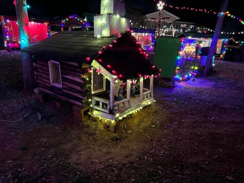 A small log cabin decorated with colorful lights, surrounded by festive decorations in a nighttime setting.
