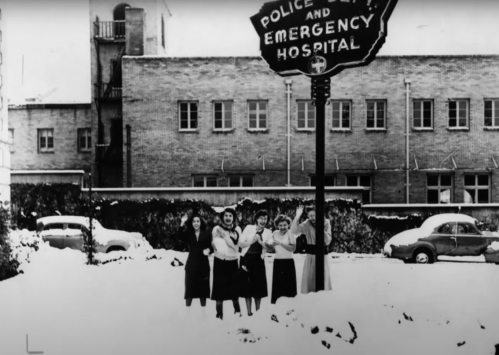 Five women stand in the snow near a sign for a police and emergency hospital, with a brick building in the background.