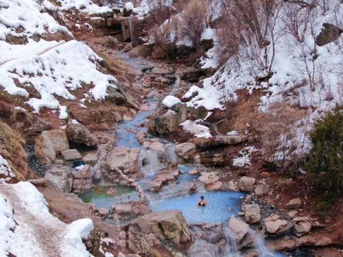 A serene hot spring surrounded by snow-covered rocks and trees, with a person relaxing in the water.