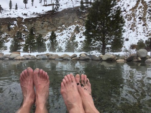 Two pairs of bare feet resting in a natural hot spring, surrounded by snow-covered rocks and trees in the background.