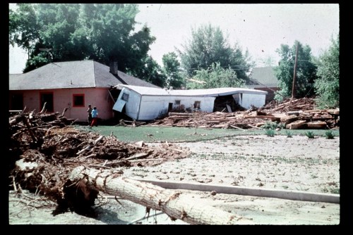 A house partially collapsed, surrounded by debris and fallen trees after a flood, with a person walking nearby.