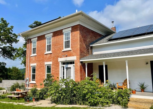 A brick house with solar panels, surrounded by greenery and a porch with plants and seating. Blue sky above.