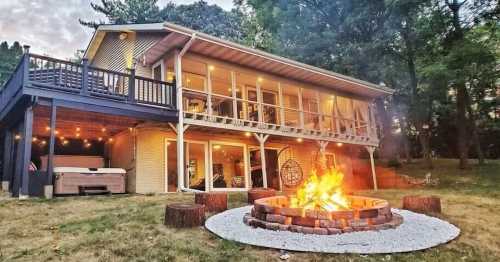 A cozy house with a fire pit in front, surrounded by trees and outdoor seating, illuminated by warm lights.