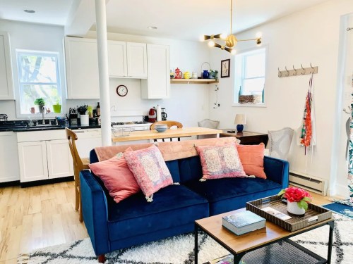 Cozy living space featuring a blue sofa with pillows, a wooden table, and a bright kitchen area in the background.