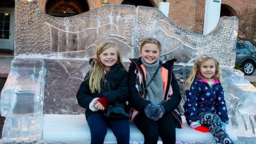 Three smiling girls sit on a large ice sculpture resembling a couch, dressed warmly for winter.