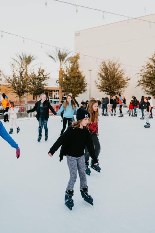 A group of people ice skating outdoors, surrounded by palm trees and festive lights.