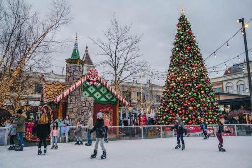 A festive outdoor ice skating rink with a decorated Christmas tree and a gingerbread house in the background.