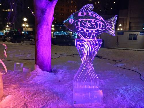 An illuminated ice sculpture of a fish, standing on a pedestal, surrounded by snow and festive lights.