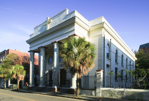 Historic building with columns and a cross, surrounded by palm trees, set against a clear blue sky.