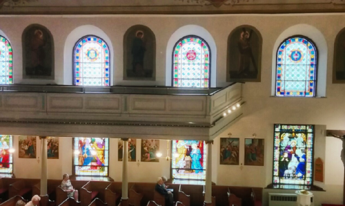 Interior of a church featuring stained glass windows, wooden pews, and religious artwork on the walls.