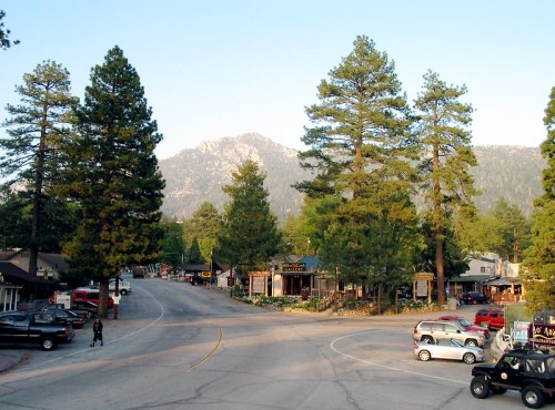A scenic view of a small town surrounded by tall pine trees and mountains, with shops and parked cars along the road.