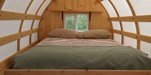 Cozy wooden interior of a small cabin with a double bed, featuring soft pillows and a green blanket.