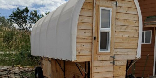A wooden trailer with a curved white roof, featuring a small window and door, set against a grassy background.